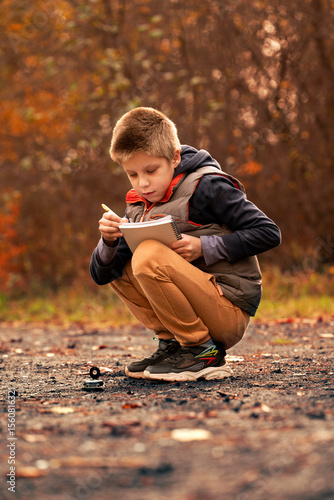 A boy on an autumn path put a compass on the ground and writes in a notebook. The atmosphere of exploration and adventure. A little pathfinder or a young tourist in an autumn forest. 