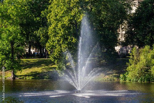 Fototapeta Naklejka Na Ścianę i Meble -  water fountain in city park with green trees and sunlight, summer nature scene with spraying water in urban garden, peaceful relaxation landscape in european city center with natural tranquility