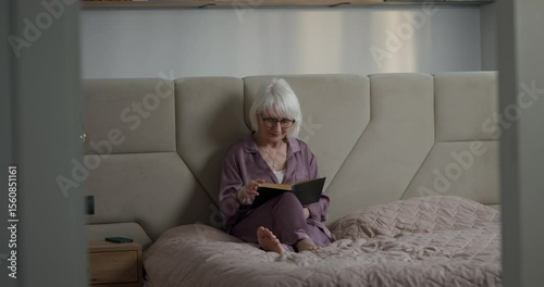 Joyful elderly woman in pajamas reading book wearing glasses sitting on bed during her leisure time during daytime. Mature woman in pajamas and glasses reading a book