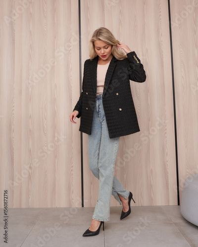 Blonde woman in stylish black blazer with gold buttons, light jeans, and black high heels stands on gray floor against wooden panel wall, looking down with hand in hair, relaxed pose