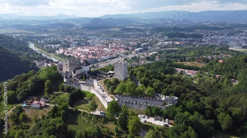 stunning aerial view of Celje Old Castle Celjski Stari Grad perched on a hill, surrounded by lush green forests. The castle overlooks the city of Celje, with the Savinja River winding through, and
