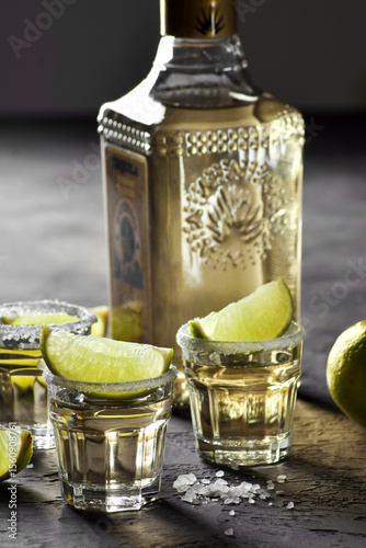 Glass shot glasses filled with golden tequila are arranged on the grey table, garnished with lime wedges and salt, creating a festive atmosphere for celebration