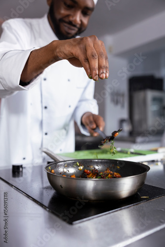 African american expert in a kitchen prepares a dish with fresh basil and parsley, showcasing culinary expertise in fine dining and food prep. Young person chef sprinkles rosemary leaves.