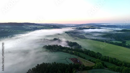 Aerial drone footage of a misty meadow surrounded by forest and mountains at sunrise. Soft golden light filters through fog, creating a tranquil, serene landscape that blends nature’s calm with early 
