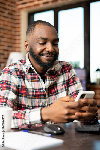 Black male entrepreneur working from home, seated at desk, checking social media on cellphone. Positive african american businessman enjoying quick chat with colleague on mobile device.