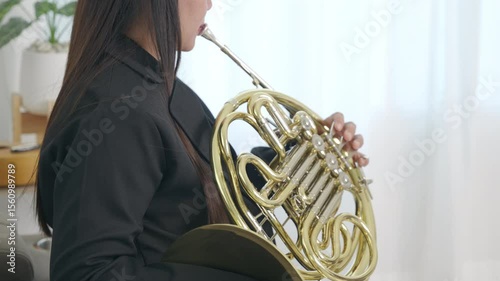 In a relaxed living room, a woman sits on a sofa, skillfully playing a shiny gold French horn. Her focus and dedication to her craft are evident in this artistic moment.