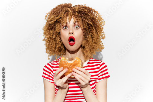 Surprised young woman with curly hair holding a burger, wearing a red and white striped shirt, expressing excitement about unhealthy food