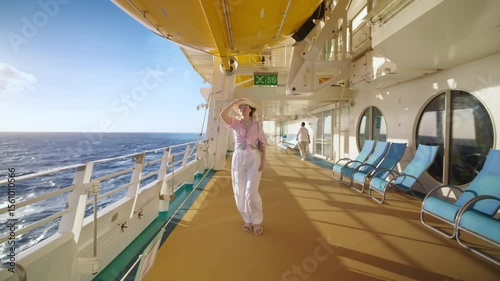 A woman is enjoying a sunny day on a luxurious cruise ship, strolling the deck, admiring the ocean views, and dressed in a chic outfit with a stylish hat, embodying elegance at sea