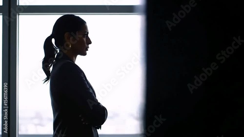 A businesswoman in formal attire standing at high office window, taking a deep breath while thinking