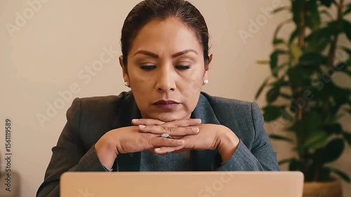 A woman in a business suit sits at a desk, laptop open, hands clasped under her chin as she stares blankly ahead, Her face shows fatigue and pressure