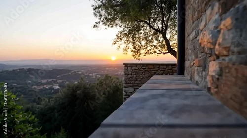 Golden sunset landscape view from building balcony featuring stone wall and tree. Soft focus on the distant hills and sky creates a peaceful atmosphere
