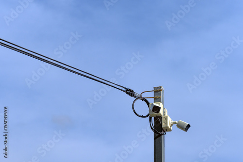 A high-angle surveillance camera mounted on a steel pole contrasts against the backdrop of a clear sky, evoking both vigilance and stillness.