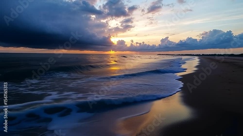 Scenic seaside view depicting a beach at sunset with golden sun reflecting in the water under dramatic cloudy sky. The wave's foam is shown in the foreground
