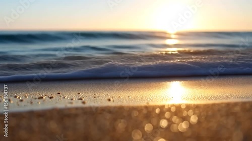 Golden hour beach scene with sun reflecting on the water surface. Focused on the sandy foreground and blurry waves. Soft lighting and warm tones dominate