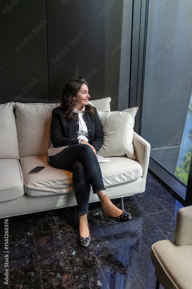Obraz premium In a modern office, a confident businesswoman relaxes on a stylish couch, embodying professionalism and sophistication while radiating positive energy that brightens the atmosphere