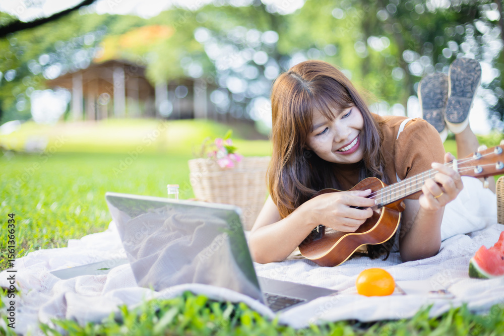 Young woman enjoys leisure time playing ukulele during picnic on grass with laptop nearby, capturing happy moment of people and family concept outdoors Young woman enjoys leisure time playing ukulele during picnic on grass with laptop nearby, capturing happy moment of people and family concept outdoors