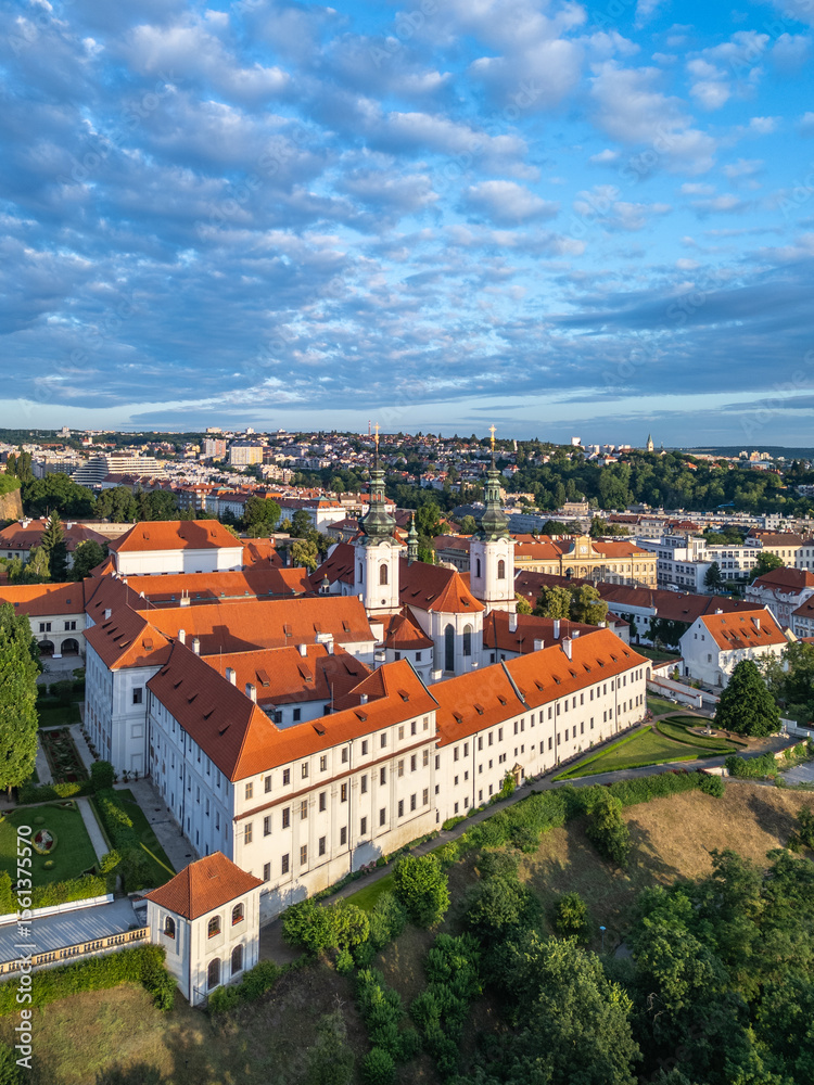 Obraz premium Strahov Monastery captures the serene beauty of Prague at sunrise, with its stunning architecture and surrounding greenery. The aerial view showcases the vibrant rooftops and peaceful atmosphere.