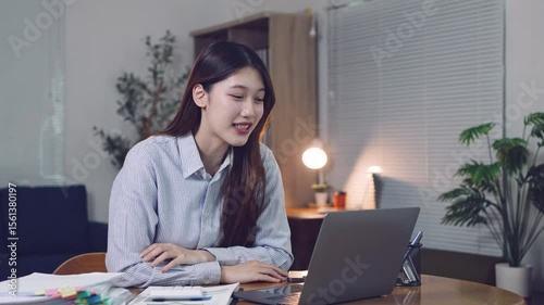 Professional asian businesswoman working late at home, video conferencing on laptop while waving and smiling during remote meeting at night