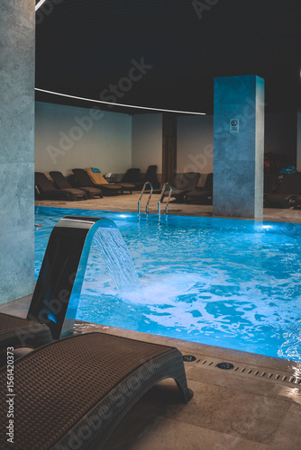 A sleek stainless fountain pours into a luminous blue indoor pool, churning bubbles beneath soft strip lights while empty loungers await in a calm, modern wellness space