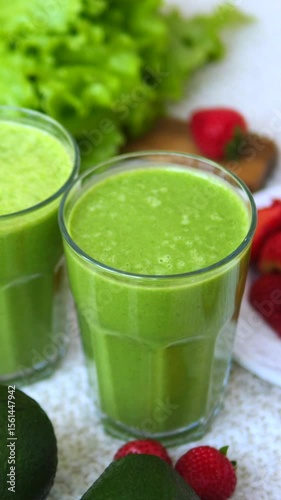 Vertical screen preparing vibrant green smoothie with fresh organic lettuce, creamy avocado, and ripe strawberries, hand carefully grasping glass against clean white background