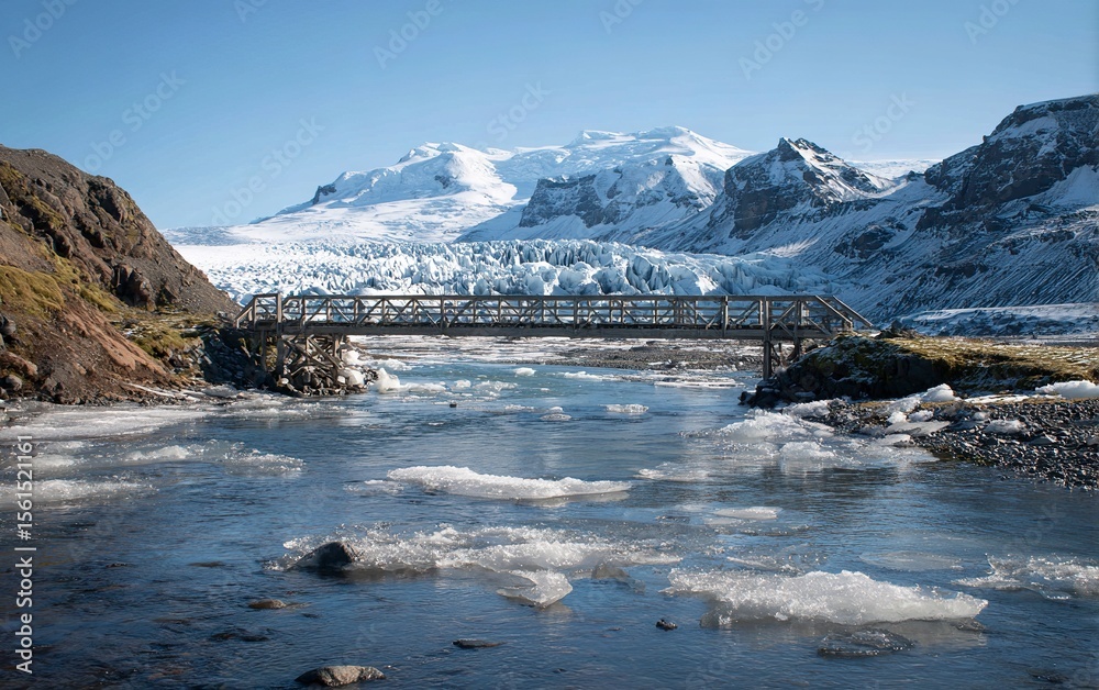 Obraz premium Photo of the Fjallabaksh Puppet Bridge in Iceland. In the background, there is an icy river and glaciers with small icebergs floating around. The photo was taken from behind the bridge,