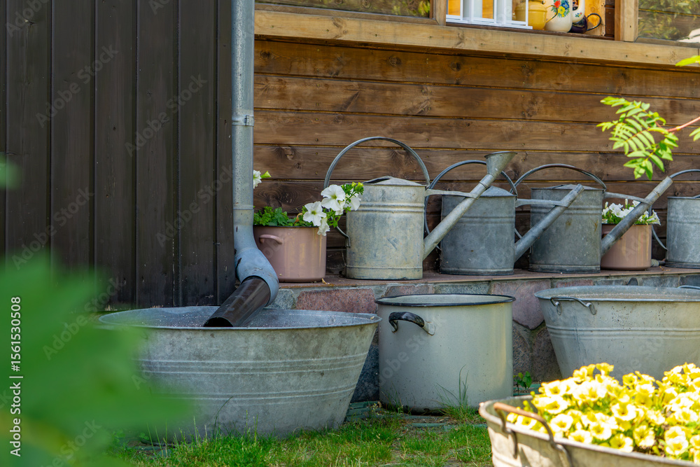 Obraz premium row of old galvanized metal watering cans and tubs placed against a wooden wall, with flower pots and blooming plants nearby. Charming rustic garden arrangement.