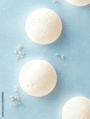 Three white spherical objects resembling moons, complete with craters, resting on a light blue surface dotted with white specks and delicate sprigs of flowers, creating a serene celestial scene
