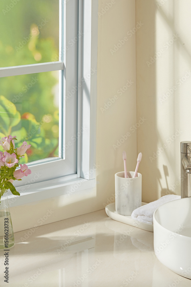 Fototapeta premium Sunlit bathroom counter with flower vase, cotton swabs, and soap bottle beside a ceramic sink. Warm tones and natural light make it ideal for wellness, hygiene, or beauty product visuals.