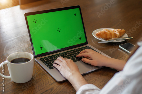 laptop blank green screen mockup. woman using laptop computer, hand typing on keyboard at home for work with hot coffee, croissant bakery and mobile phone on the wooden table in the morning.