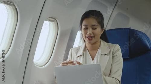 Professional asian businesswoman conducting video conference while working remotely during international flight, utilizing laptop with wireless connectivity in airplane cabin