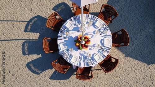 aerial view of round dining table, Top view of round outdoor table set for eight with white tablecloth, cutlery, glasses, and fruit centerpiece. Surrounded by wicker chairs and shadows