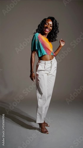 Smiling Black woman poses confidently in color-blocked top and white high-waisted pants in studio. Bright lighting and modern styling. Concept: fashion confidence, joyful personality