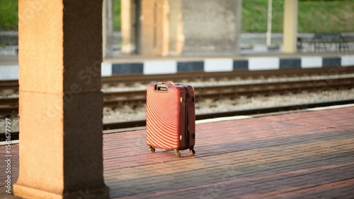 A red hard-shell suitcase with wheels and a textured surface stands upright on a brick train station platform in direct sunlight, with railway tracks and platform columns in the background.