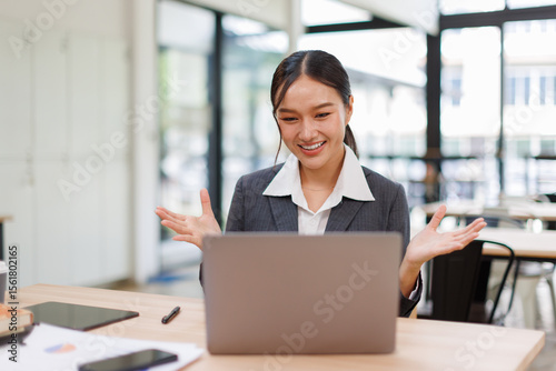 Excited friendly young asian woman 20s in casual clothes sit on desk,  talk work on laptop pc computer get video call gesticulate in living room at workplace.
