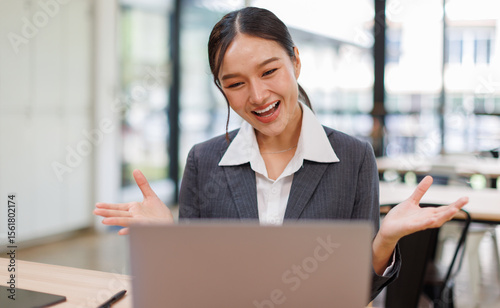Excited friendly young asian woman 20s in casual clothes sit on desk,  talk work on laptop pc computer get video call gesticulate in living room at workplace.
