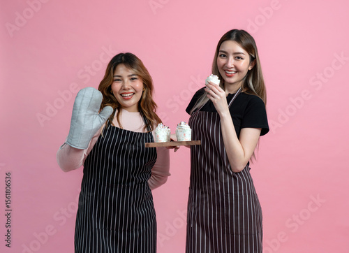 Young asian couple with apron in celebratory or festive mood holding a tray with cupcake, all decorated with white frosting and colorful sprinkles. Portrait on pink background with studio light.