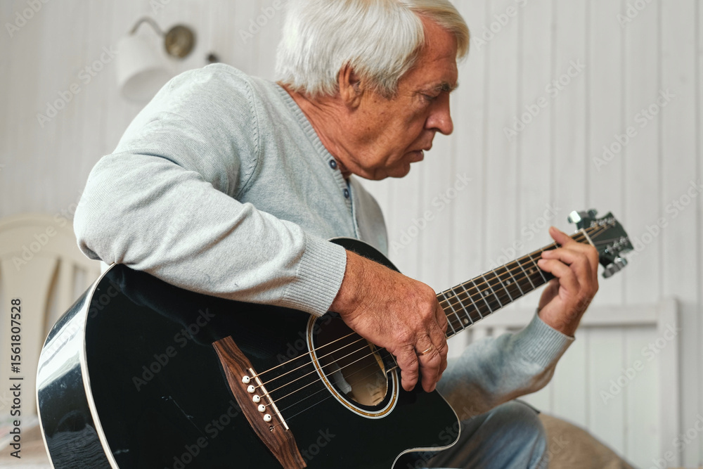 Obraz premium Senior man is playing guitar. Elderly male sitting at sofa and play music. Portrait of a gray haired mature 60s person with acoustic guitar, he is learning to play. Enjoying retirement life at home.