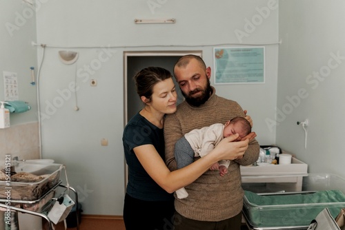 A couple lovingly holds their newborn baby in a cozy indoor setting. The mother gazes at the baby while the father supports them, showcasing a moment of tenderness and family bonding.