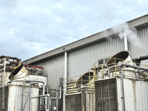 Industrial cooling tower system emitting vapor under blue sky. Heavy-duty piping, valves, and heat exchangers in a manufacturing or refinery plant exterior.