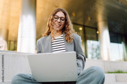 Free Download Stylish woman with laptop sitting on steps against modern building background. Business woman in glasses working outdoors on laptop. Freelance, business and break concept. - GFXLoad