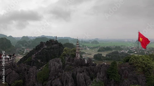 Aerial drone footage of a Vietnamese flag waving in the wind on Hang Múa viewpoint, with a stone pagoda and lush green landscapes in Ninh Binh, Vietnam