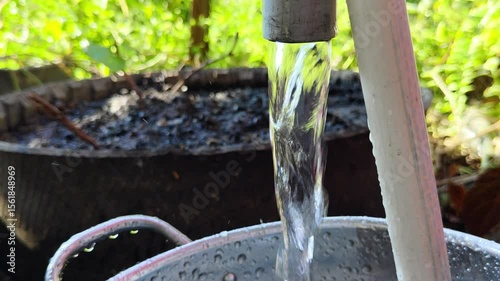 A stream of water flowing from a tap into a metal container, in an outdoor garden setting, showcases hydration and access to clean water.