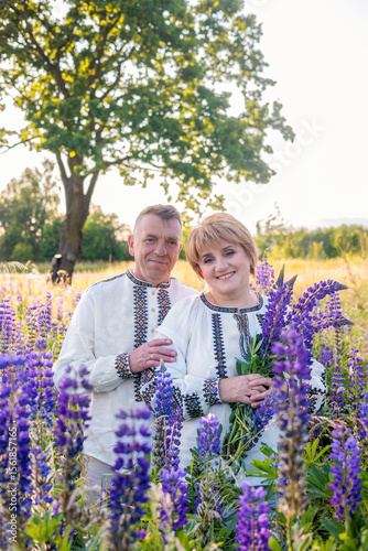 Smiling mature Ukrainian couple wearing vyshyvanka shirts and holding lupine flowers, standing outdoors in a rural landscape. Concept of love, tradition, family unity, countryside lifestyle.

