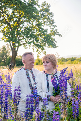 Smiling mature Ukrainian couple wearing vyshyvanka shirts and holding lupine flowers, standing outdoors in a rural landscape. Concept of love, tradition, family unity, countryside lifestyle.

