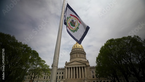 The West Virginia state capitol building features its iconic dome and stone facade, with a flag waving in front amidst trees under an overcast sky.