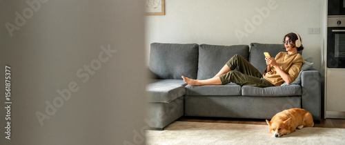 Smiling young student woman lying on couch, listening music in headphones on mobile phone application. Relaxing girl at new real estate apartment home using mobile phone online app. Banner, copy space