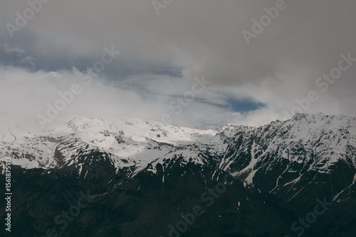 View of the snow-covered peaks of the Caucasus Mountains. The sun is covered by clouds. It is cloudy