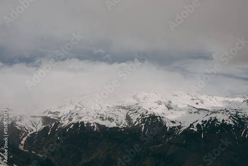 View of the snow-covered peaks of the Caucasus Mountains. The sun is covered by clouds. It is cloudy