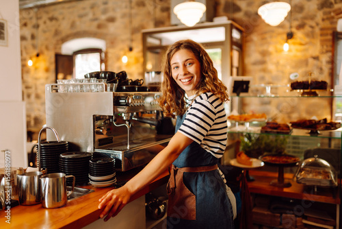 Happy female barista holding a cup of hot and aromatic coffee in her hands standing behind the bar. Young employee of the coffee shop gives coffee to a customer. Concept of drinks, small business.