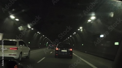 Point of view from inside a car driving through a long, illuminated tunnel at night. The scene shows a car ahead and the rhythmic pattern of tunnel lights, evoking movement, focus, and modern